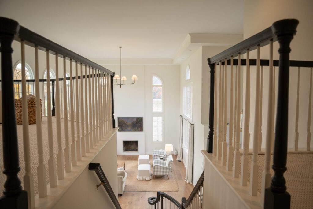 View from staircase landing overlooking a living room with tall windows, a fireplace, armchairs, TV, and light neutral decor—showcasing expert interior painting in Alto Michigan. - Jeff Bryant Painting