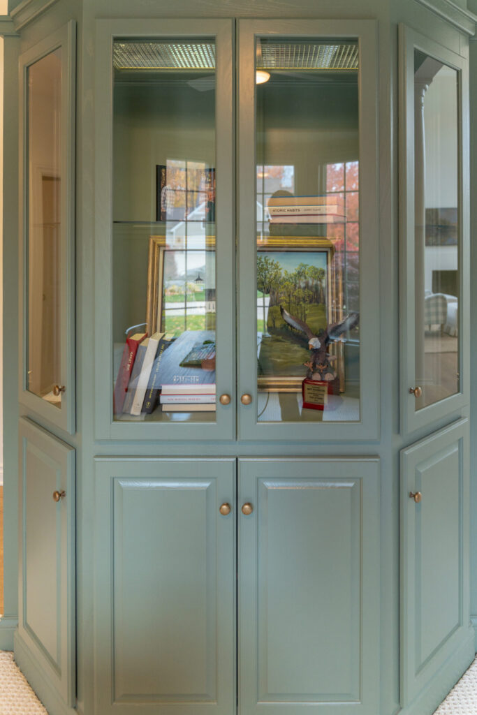 Green glass-front cabinet with books, framed photos, and a small bird figurine displayed on shelves; a tasteful addition to any space inspired by interior painting trends in Alto Michigan. Lower section has closed doors. - Jeff Bryant Painting