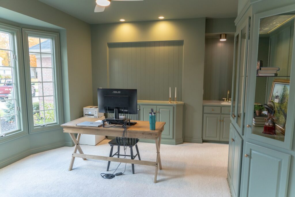 A home office with a wooden desk, computer monitor, chair, and built-in cabinets showcases fresh interior painting. Sunlight streams through windows, and there’s a small sink area in the corner—perfect for a workspace in Alto, Michigan. - Jeff Bryant Painting