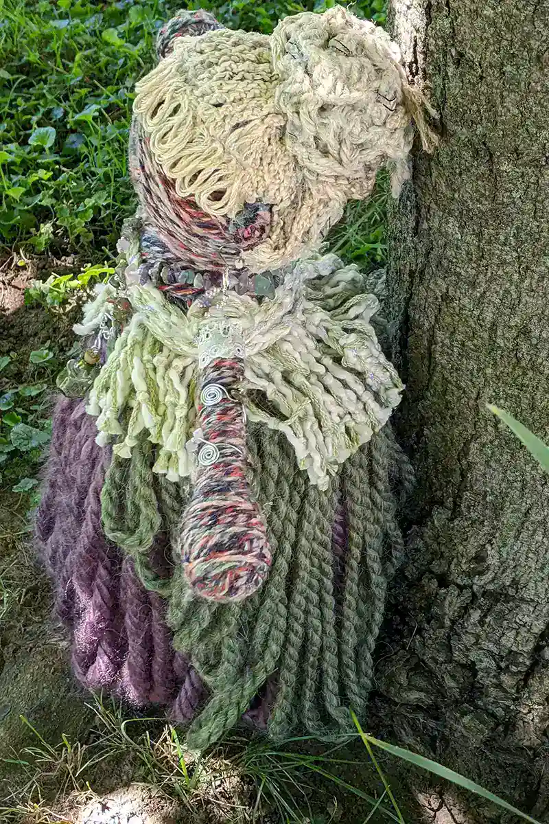 Side view of Leela the health and wellness art doll standing by a tree, with her intricate yarn body, gemstone necklace, and spiral accessories visible.