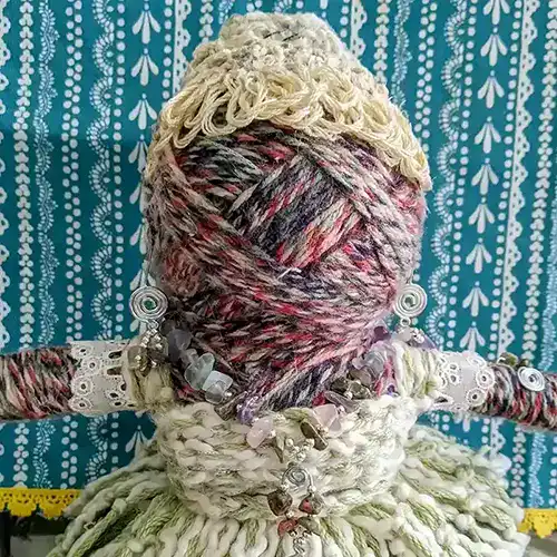 Front view of Leela the health and wellness art doll, showing her face wrapped in multicolored yarn and her "lunatic fringe" bangs, complemented by a gemstone necklace and spiral earrings.