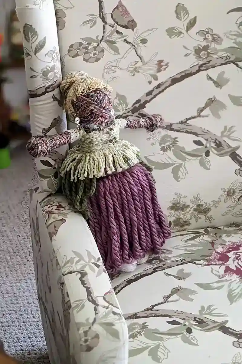 Leela the health and wellness art doll, sitting in a floral-patterned vintage armchair, showcasing her handcrafted details with a relaxed front view.