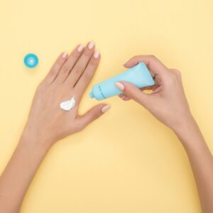 Grande plano das mãos de uma mulher a aplicar creme sobre um fundo amarelo pastel. Ideal para temas de beleza e cuidados com a pele.