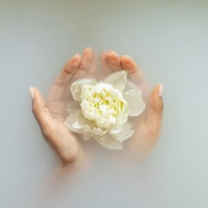 Mãos a segurar uma flor branca numa água leitosa serena, perfeita para temas de spa e bem-estar.