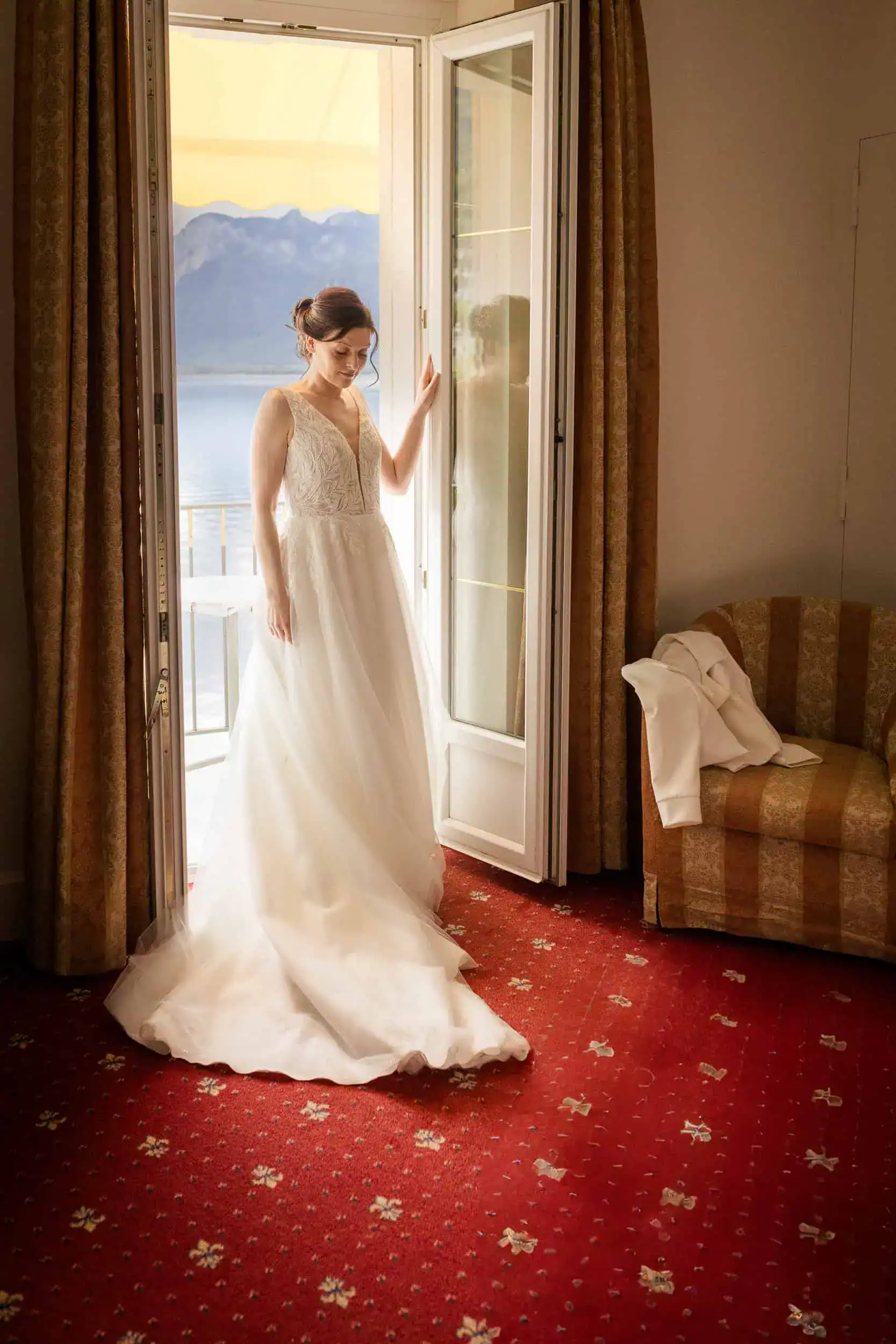 Braut im Hochzeitskleid steht träumend am Fenster mit Seeblick