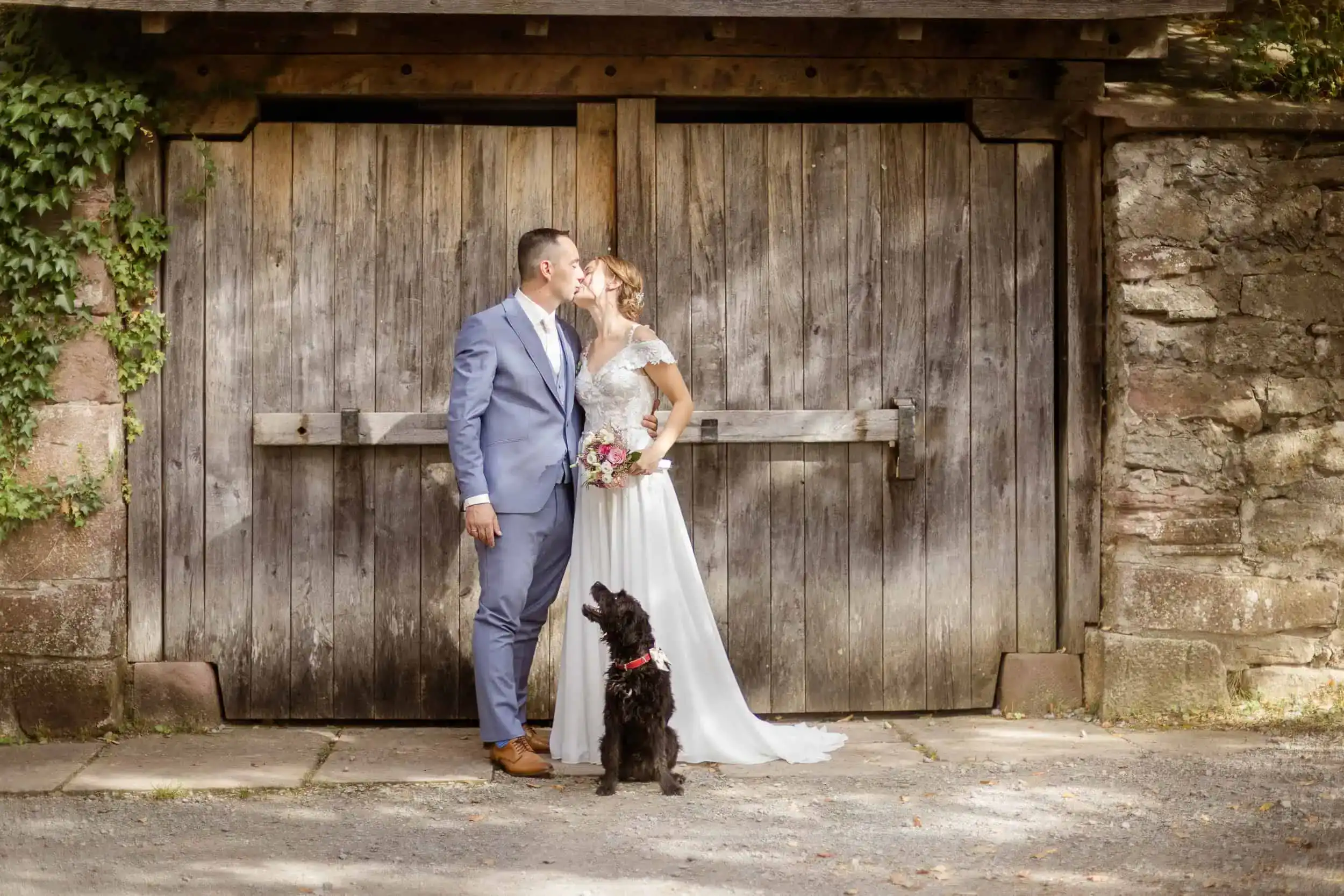 Brautpaar küsst sich vor Holztor mit Hund bei rustikaler Landhochzeit