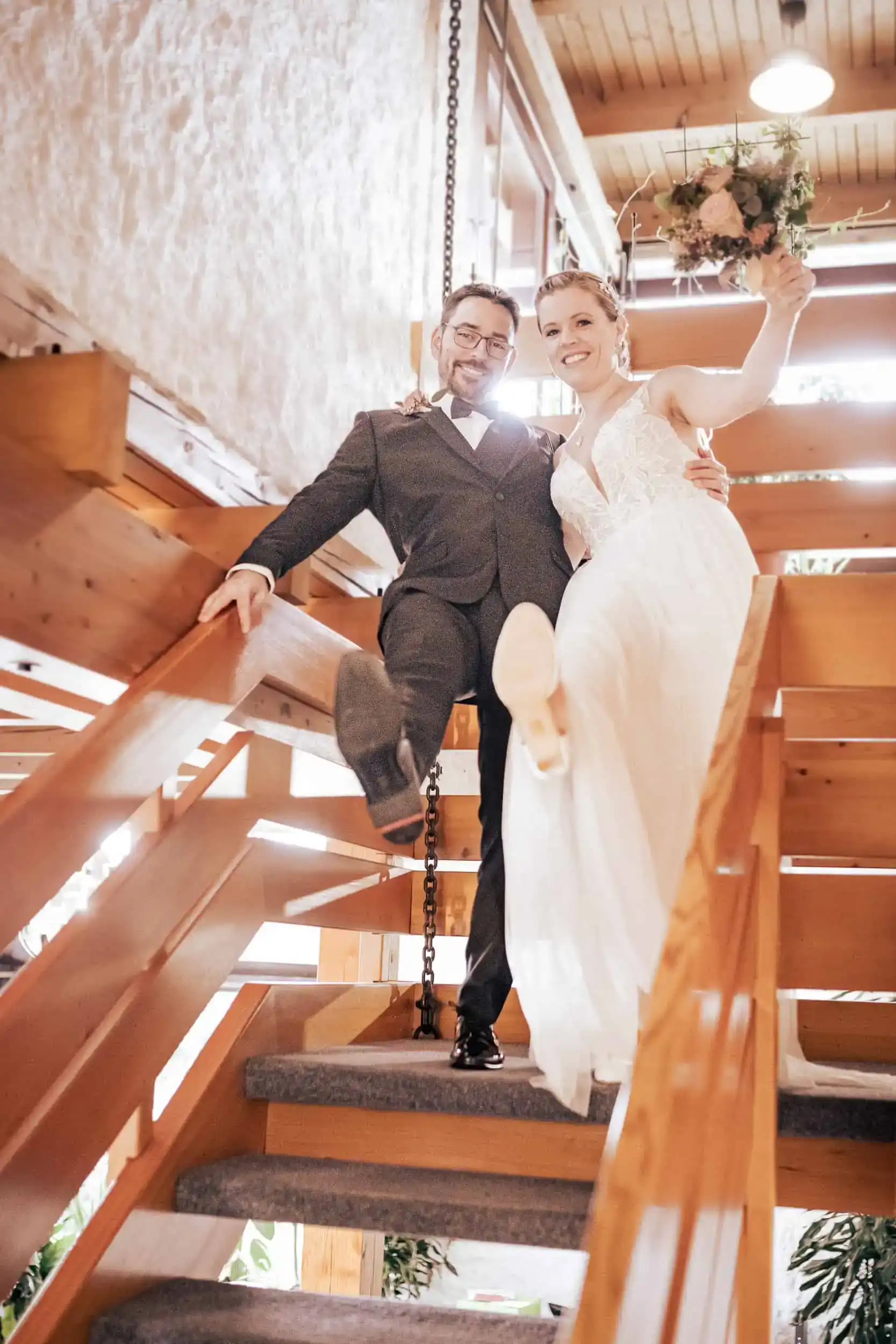 Lachendes Brautpaar auf Holztreppe mit Blumenstrauß, fröhlicher Hochzeitsmoment