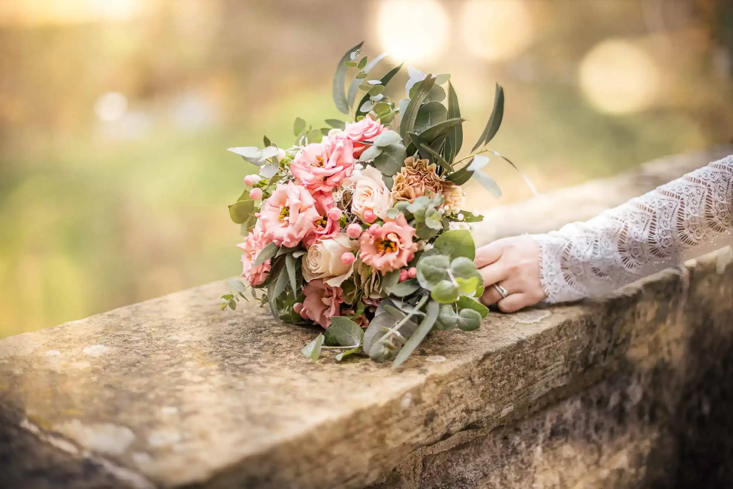 Brautstrauß mit Rosen auf Mauer, Hand der Braut mit Ring in Spitzenkleid sichtbar