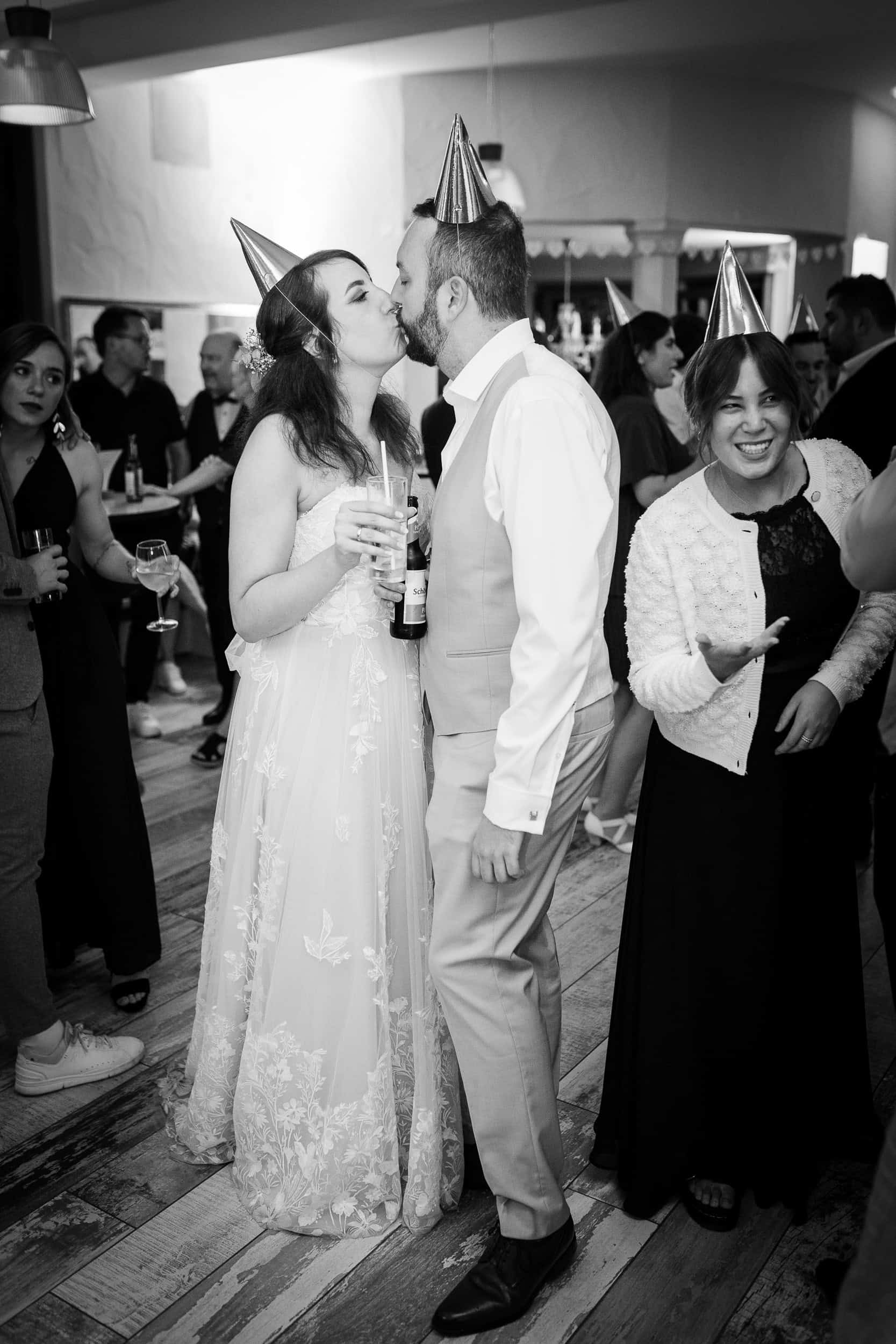 hochzeitsfotograf Pforzheim Hochzeitspaar küsst sich bei Feier mit Partyhüten, umgeben von Freunden und Familie, festliche Stimm.