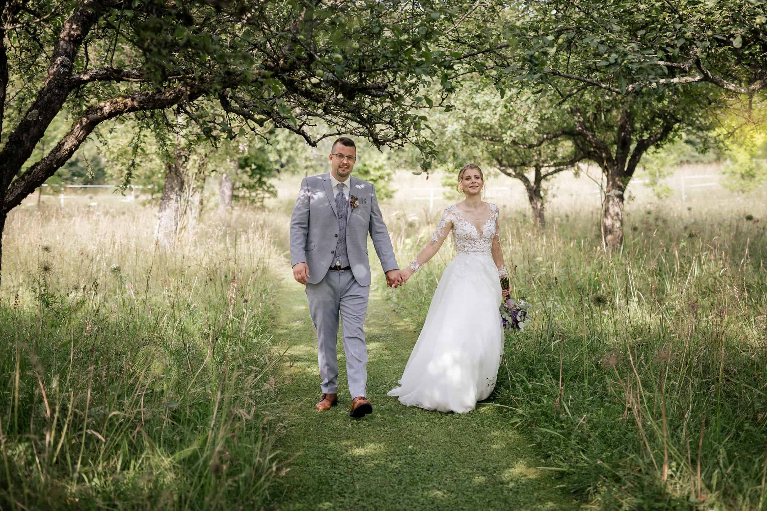 Brautpaar spaziert Hand in Hand durch blühende Obstwiese bei ihrer Sommerhochzeit.
