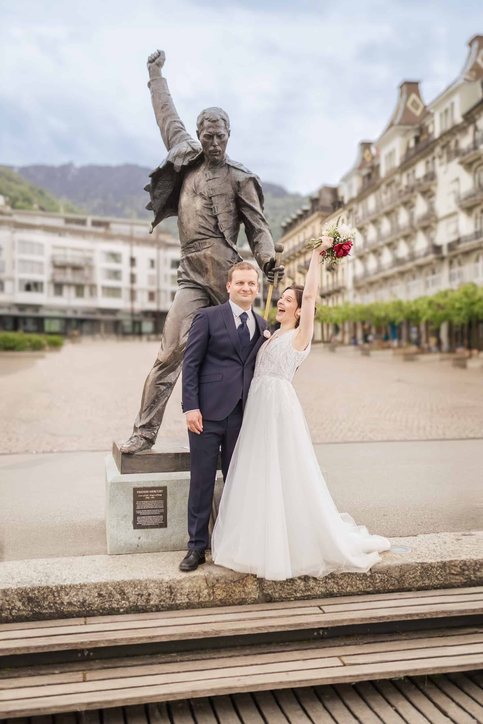 Brautpaar posiert fröhlich vor Freddie-Mercury-Statue bei Hochzeit in Montreux.