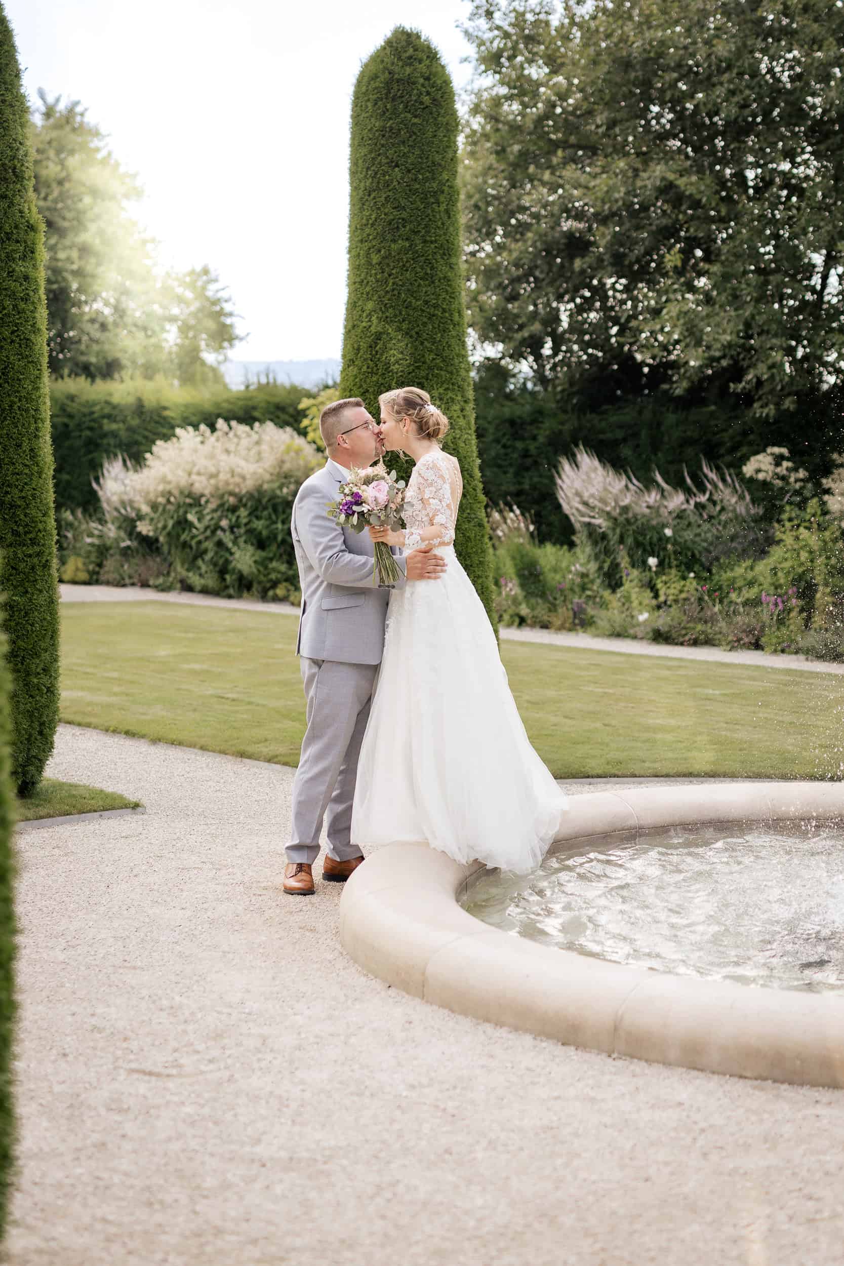 Hochzeitspaar im Garten vor Brunnen, umgeben von gepflegten Bäumen und Blumen.