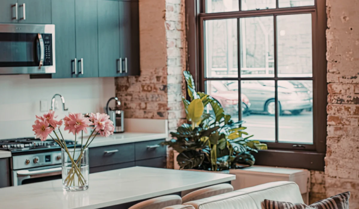 Kitchen with exposed interior brick wall, dark cabinets, large window and countertop with a vase of pink flowers