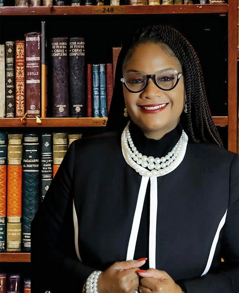 Sonya Jones A woman wearing glasses, a black and white outfit, and pearl jewelry stands smiling in front of a bookshelf filled with old books.