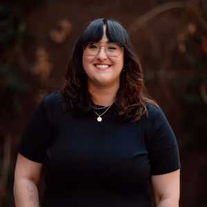 Our Team 12 Happy woman with dark hair, bangs, glasses, black shirt, and two necklaces.