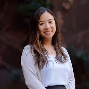Our Team 2 Smiling young Asian woman with long wavy brown hair wearing a white top and cardigan.