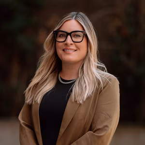 Our Team 6 Smiling blonde woman wearing black glasses, a silver necklace, and a brown blazer.