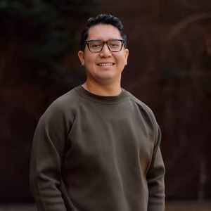 Our Team 10 Smiling man with dark hair, glasses, and a dark green sweater. Blurred background.