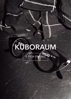 kuboraum-final1_3 Monochromatic image of black glasses on a dark, scratched surface, beside plaid fabric.
