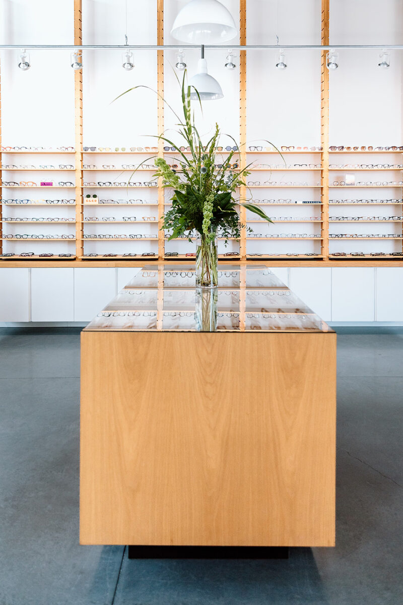 SPECTACLE - Modern Vision Care + Optical Modern optical shop with central wooden display, flowers, and numerous glasses on wall shelves.
