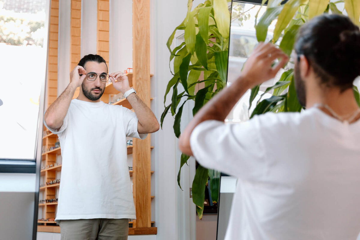 SPECTACLE - Modern Vision Care + Optical Bearded man trying on glasses, intently viewing his reflection in an optician's mirror.