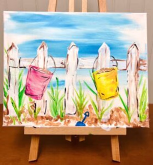 A painting on an easel depicts a white fence with a pink and a yellow bucket hanging on it, green grass, brown sand, and a blue sky in the background. - Spruced Studio Upscale Workshop