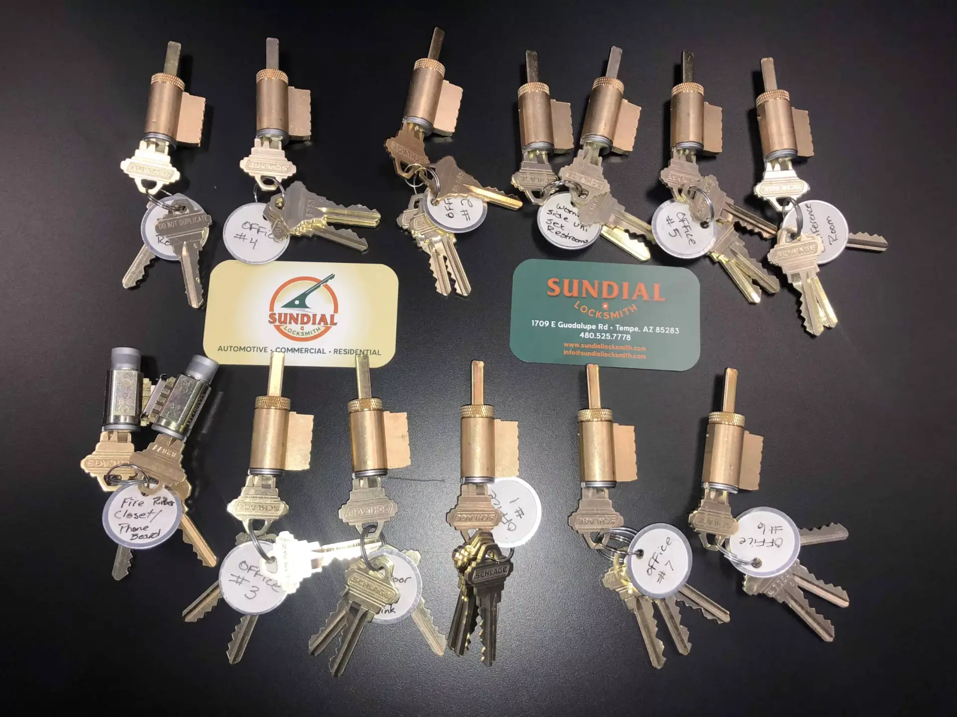 Rekey or Replace Locks A large collection of brass lock cylinders each with multiple keys displayed on a dark surface alongside Sundial Locksmith business cards.