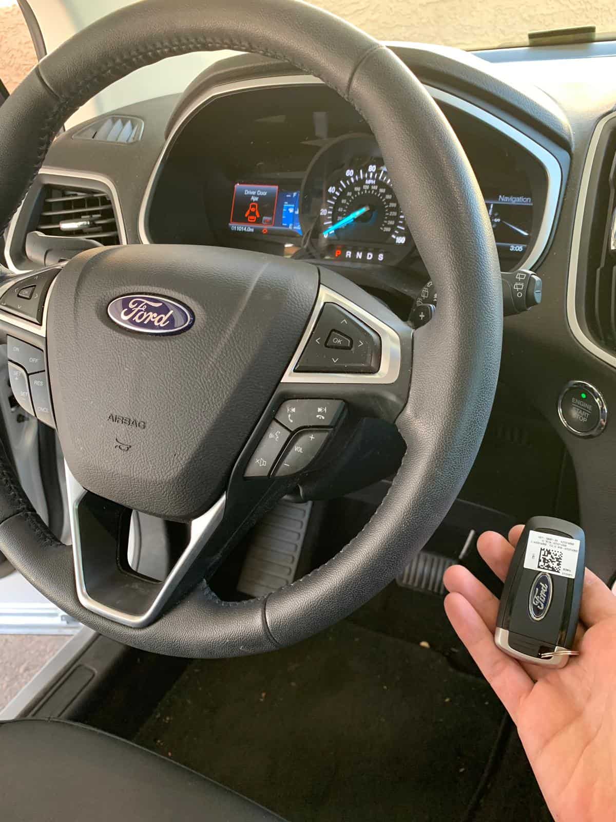 Automotive Smart Key Duplication & Programming A Ford smart key fob held near the steering wheel and dashboard of a Ford vehicle, showing the interior controls and instrument cluster.
