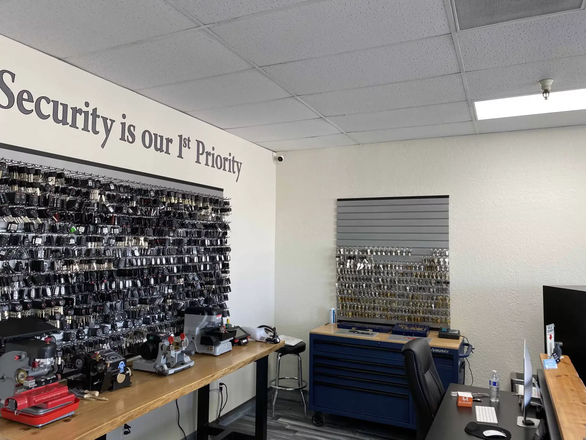 WHERE TO GET KEYS MADE? A locksmith shop interior featuring a large wall display of locks and hardware, a key cutting rack, workbench with tools, and a sign reading 'Security is our 1st Priority'.