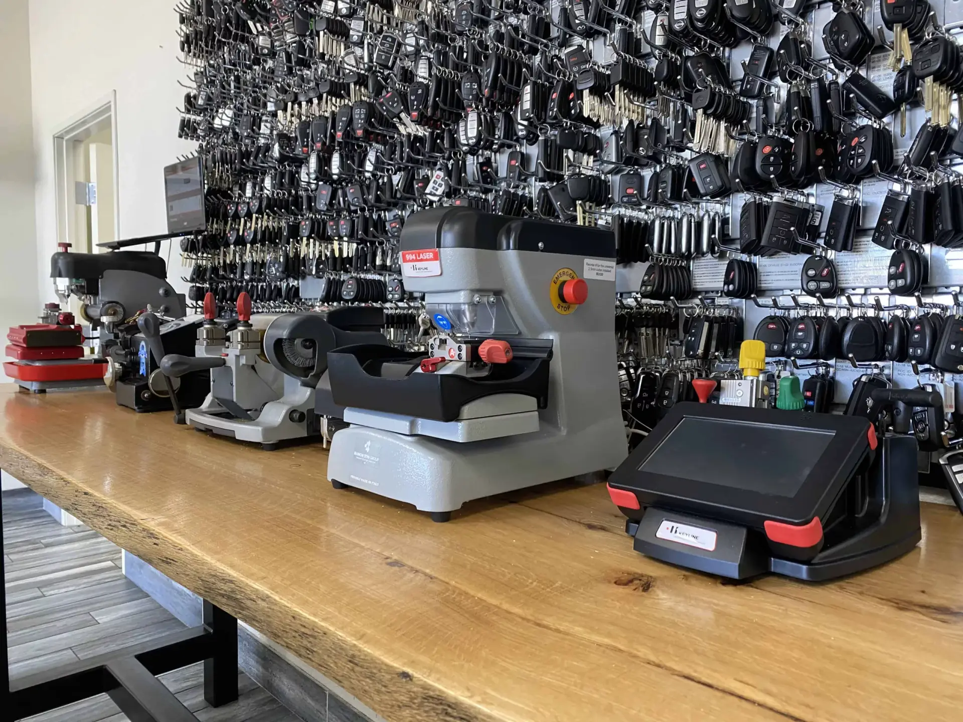 KEY DUPLICATION NEAR ME Multiple key cutting and duplicating machines lined up on a wooden workbench in front of a wall display filled with hundreds of key blanks and fobs.