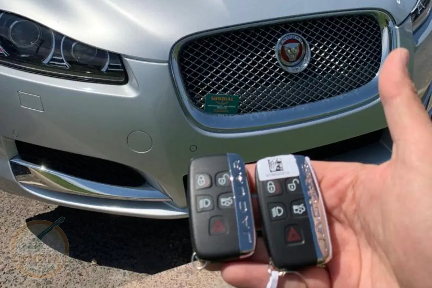 Jaguar Key Fob Replacement Cost A hand holding two Jaguar smart key fobs in front of a silver Jaguar vehicle in a parking lot.
