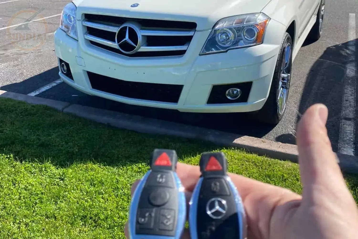 MERCEDES BENZ KEY REPLACEMENT A hand holding two Mercedes-Benz smart key fobs in front of a white Mercedes-Benz GLK SUV parked in a lot.