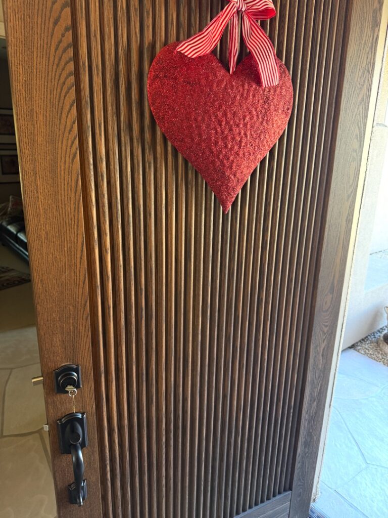 How a Locksmith Can Walk Through Your Home and Tell You What's Weak A brown wooden front door with vertical grooves, featuring a red fabric heart decoration hanging from a red ribbon at the top.