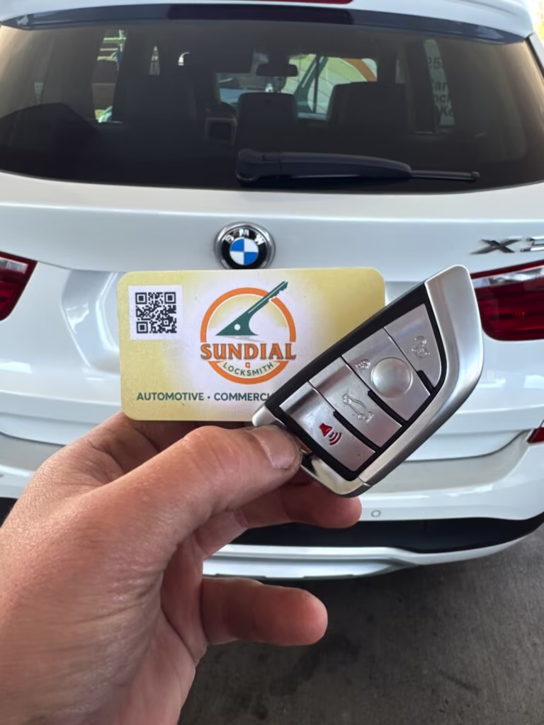 BMW X3 Key Fob Replacement Service A hand holding a BMW smart key fob in front of a white BMW X3 SUV with a Sundial Locksmith business card.