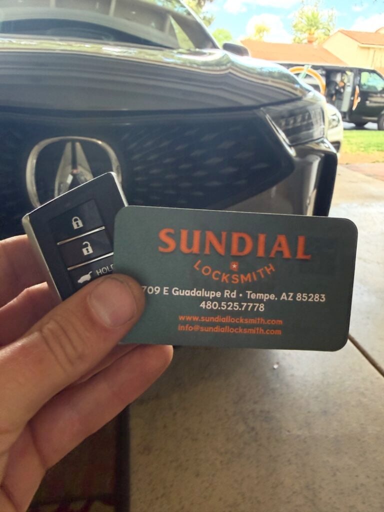 Rekey or Replace? How to Decide Without Overspending A hand holding a modern smart car key fob next to a Sundial Locksmith business card in front of a parked vehicle.