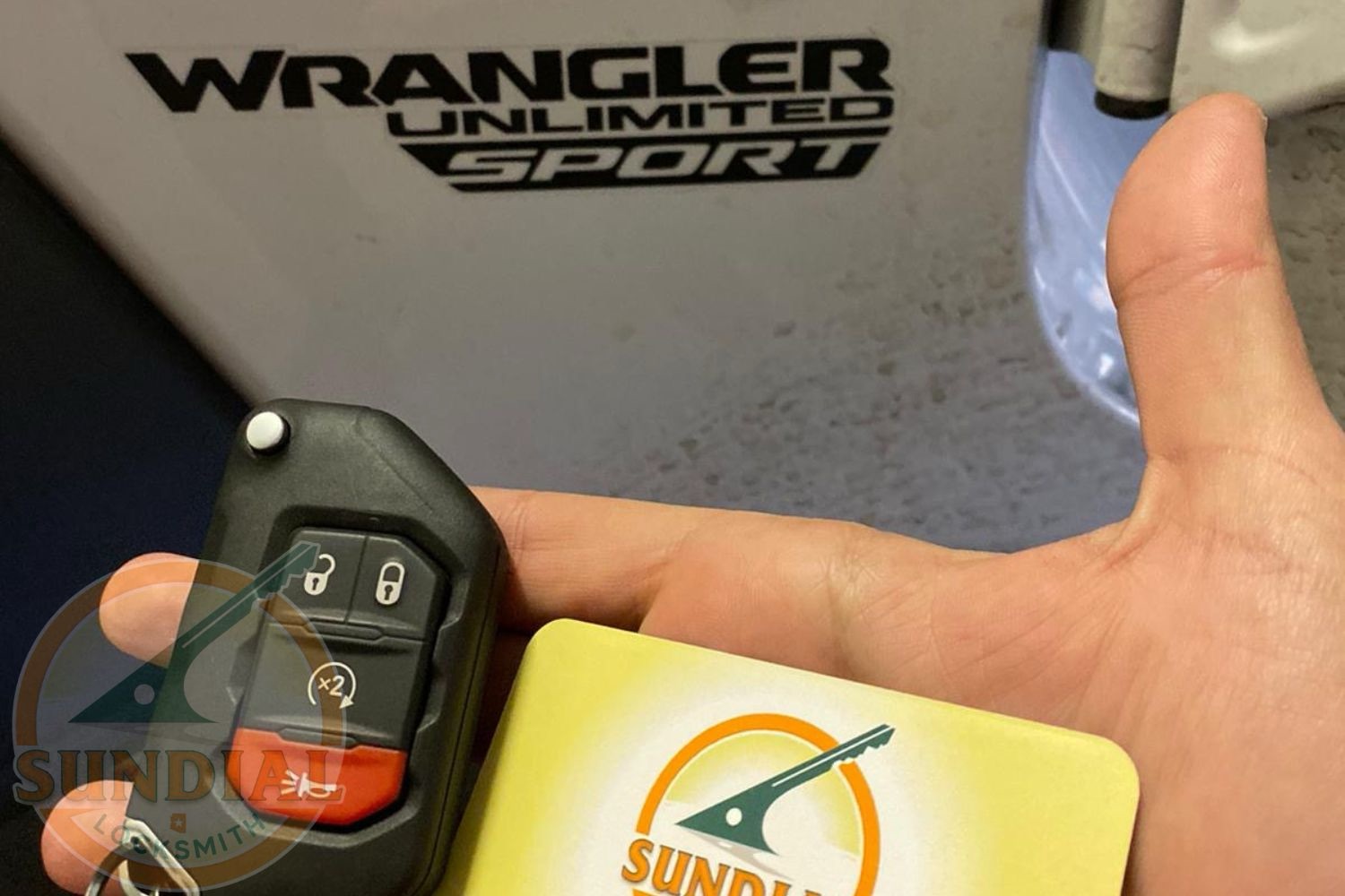 Jeep Wrangler Key Replacement: What Off-Road Owners Need to Know A hand holding a Jeep Wrangler key fob next to a Sundial Locksmith business card in front of a Jeep Wrangler Unlimited Sport vehicle.