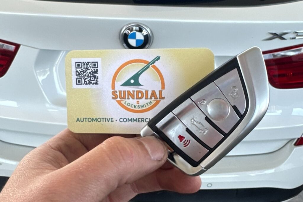 Rekey or Replace? How to Decide Without Overspending A hand holding a BMW smart key fob next to a Sundial Locksmith business card in front of a white BMW SUV.