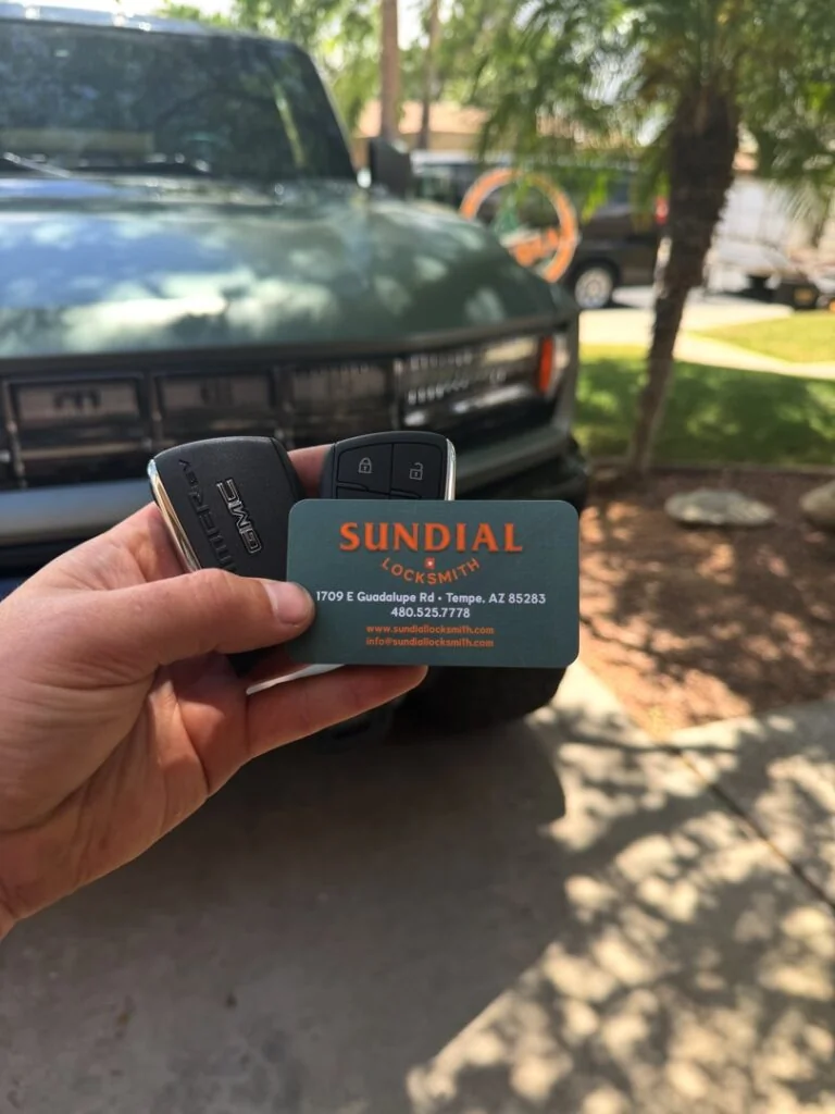 Can You Drive with a Bad Ignition Cylinder — or Is That a Bad Idea? A hand holding a Sundial Locksmith business card alongside a black car key fob, with a green truck parked in a driveway in the background.