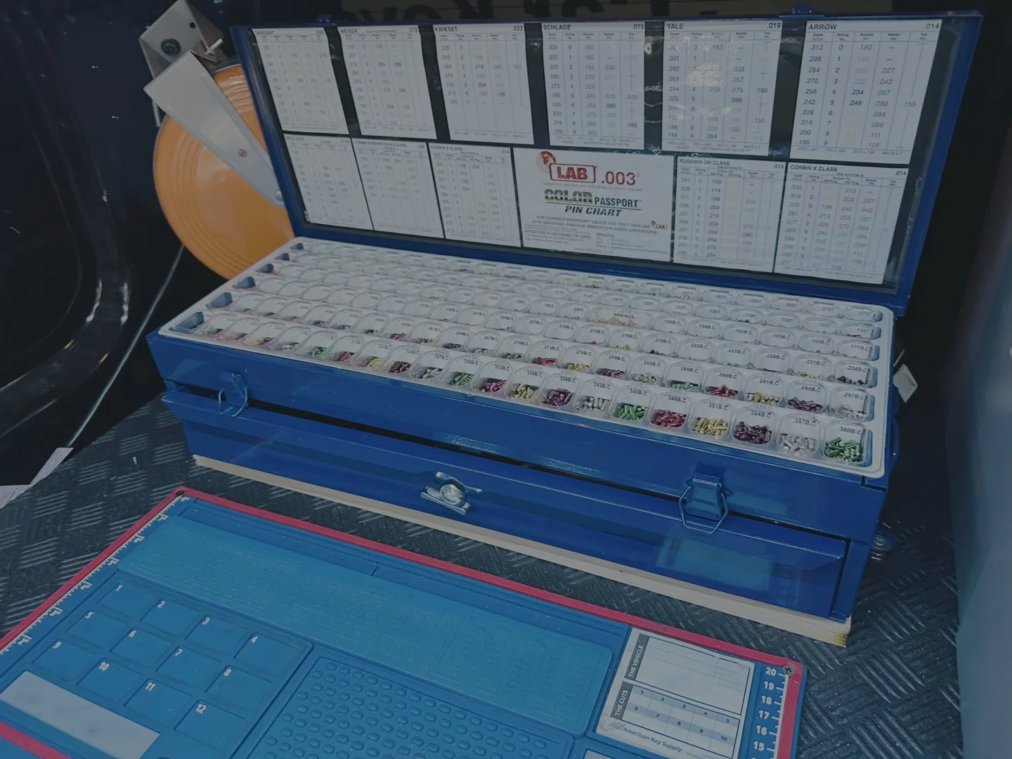 How to Get the Key Code for Your Car — and What a Locksmith Does with It A blue locksmith case open in a vehicle trunk, filled with rows of key blanks and code reference charts, alongside a blue laptop.