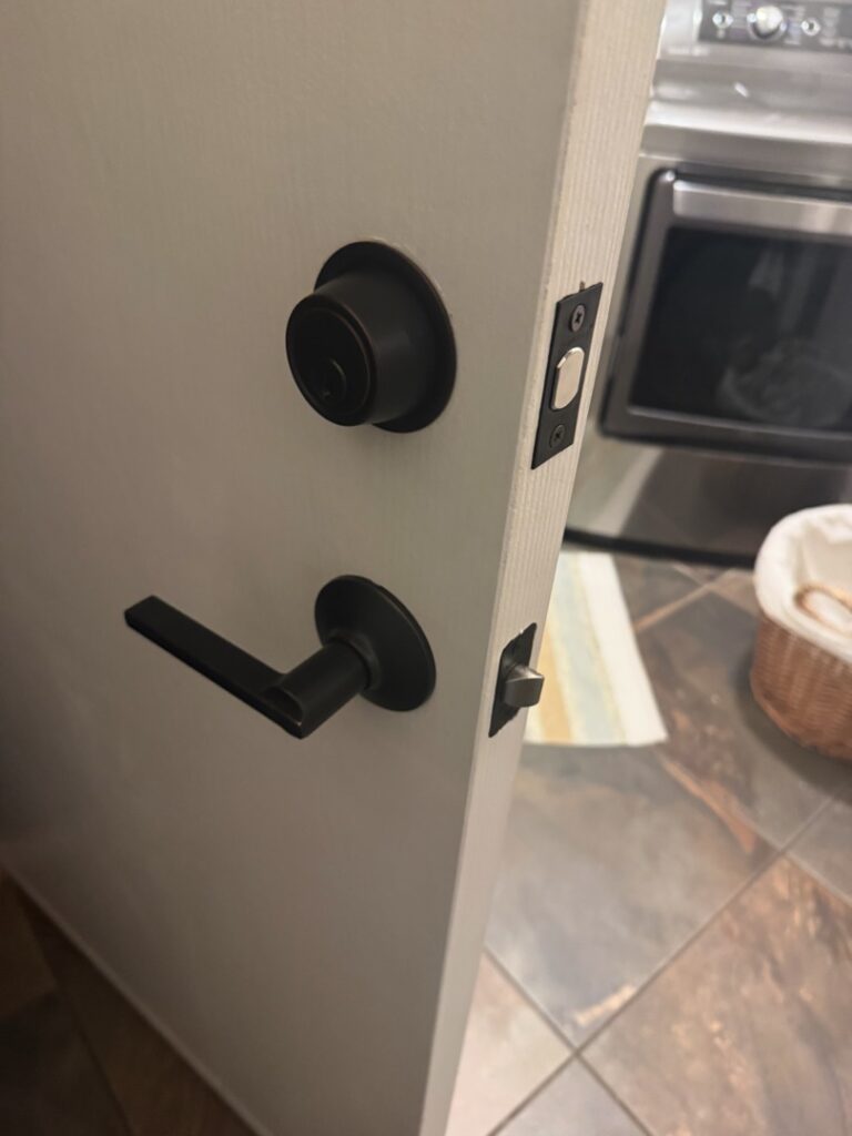 Black Door Knob and Lever Handle Installation A residential interior door featuring a newly installed matte black deadbolt and lever-style door handle set on a white door frame with tile flooring visible in the background.