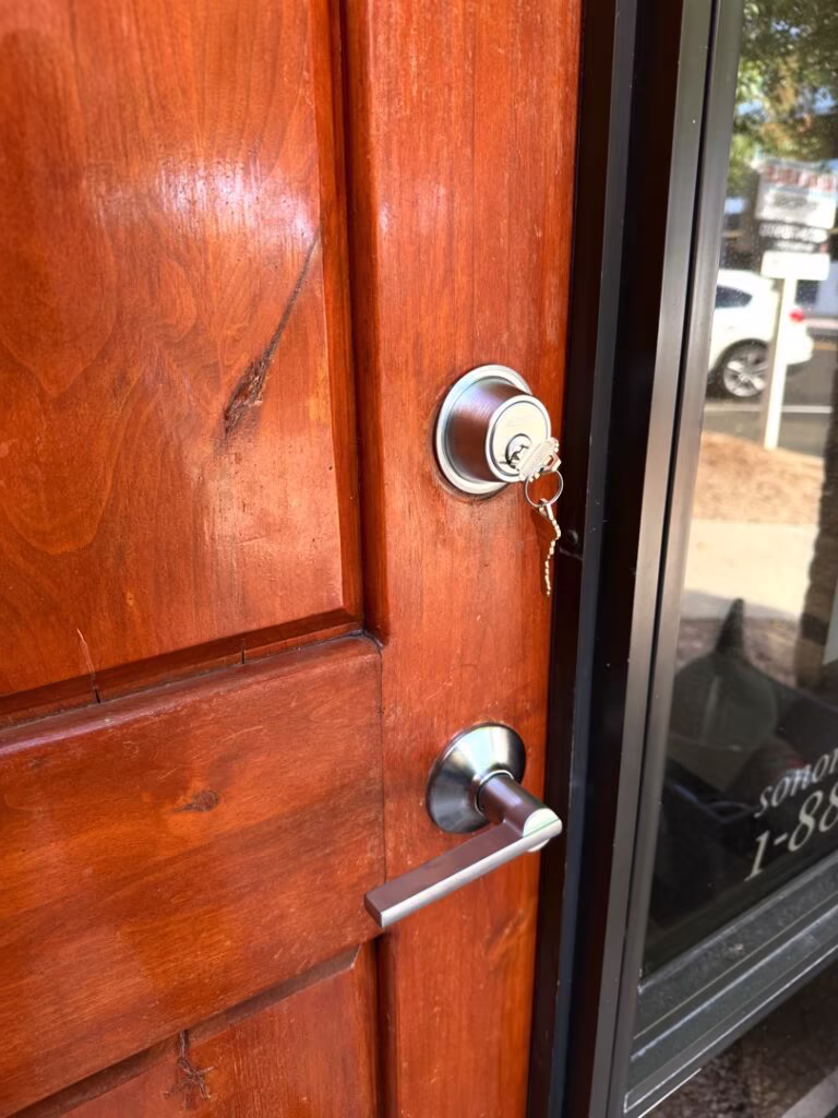 Dead Bolt Lock Installed on Wooden Door A silver deadbolt lock with keys inserted sits above a lever handle on a solid wood entry door with a glass sidelight panel.