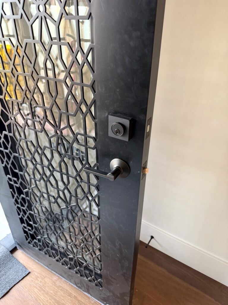 Grade 1 Deadbolt Installation: What It Is and Who Actually Needs One A dark metal security door featuring an ornate geometric lattice pattern, fitted with a satin nickel deadbolt and door knob hardware on a residential entryway.