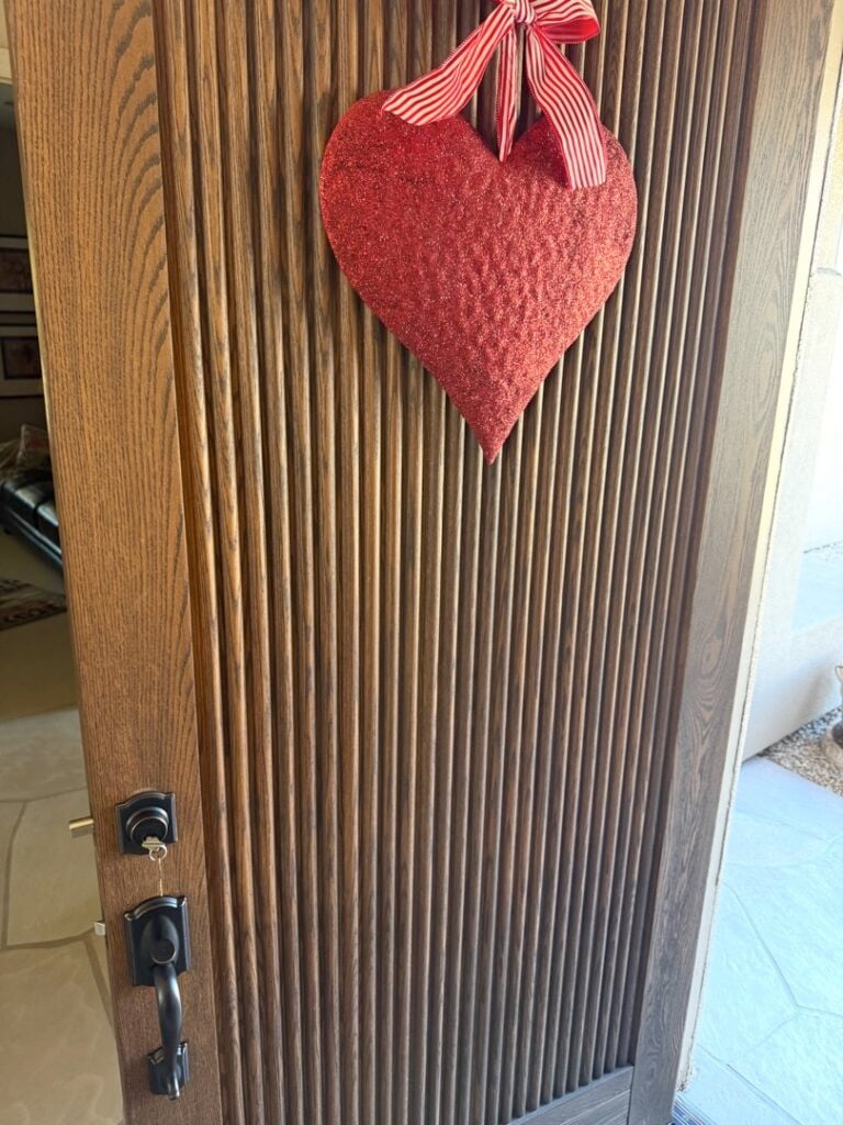 What Makes a Lock 'High Security' — and Is It Worth the Extra Cost? A wood-paneled front door with a black door handle and lock, decorated with a hanging red fabric heart ornament tied with a red ribbon.
