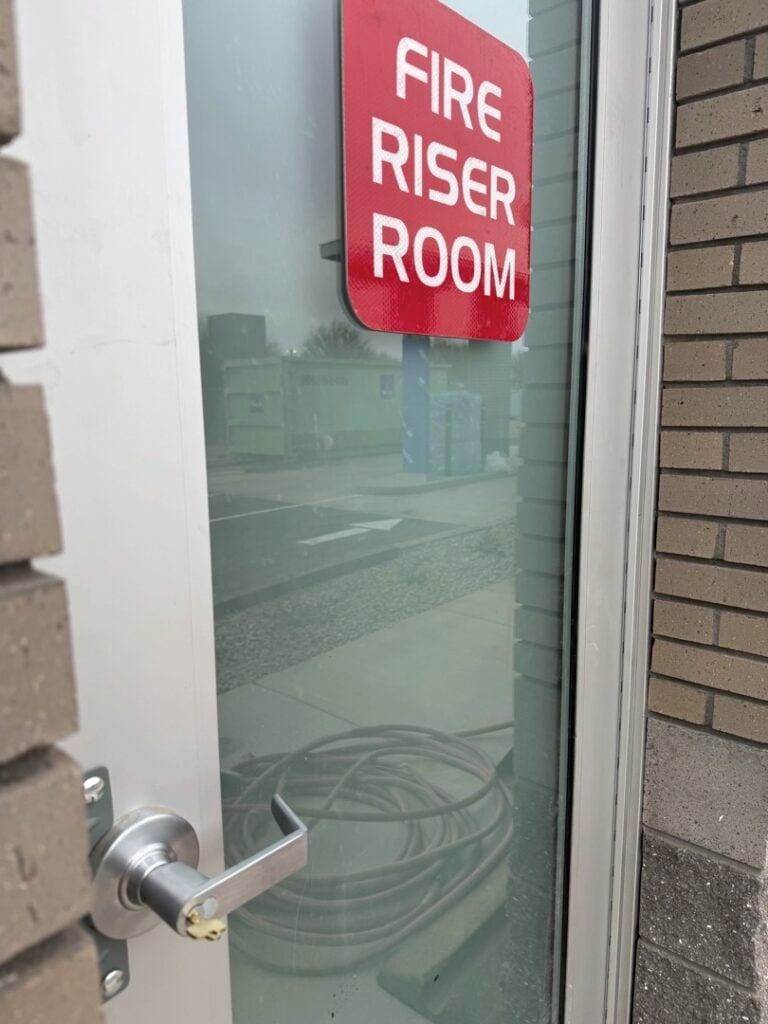 Access Control for an Office Suite: What to Expect from Installation A glass-panel commercial door with a red Fire Riser Room sign and a silver lever handle, located at the entrance to a building utility room.