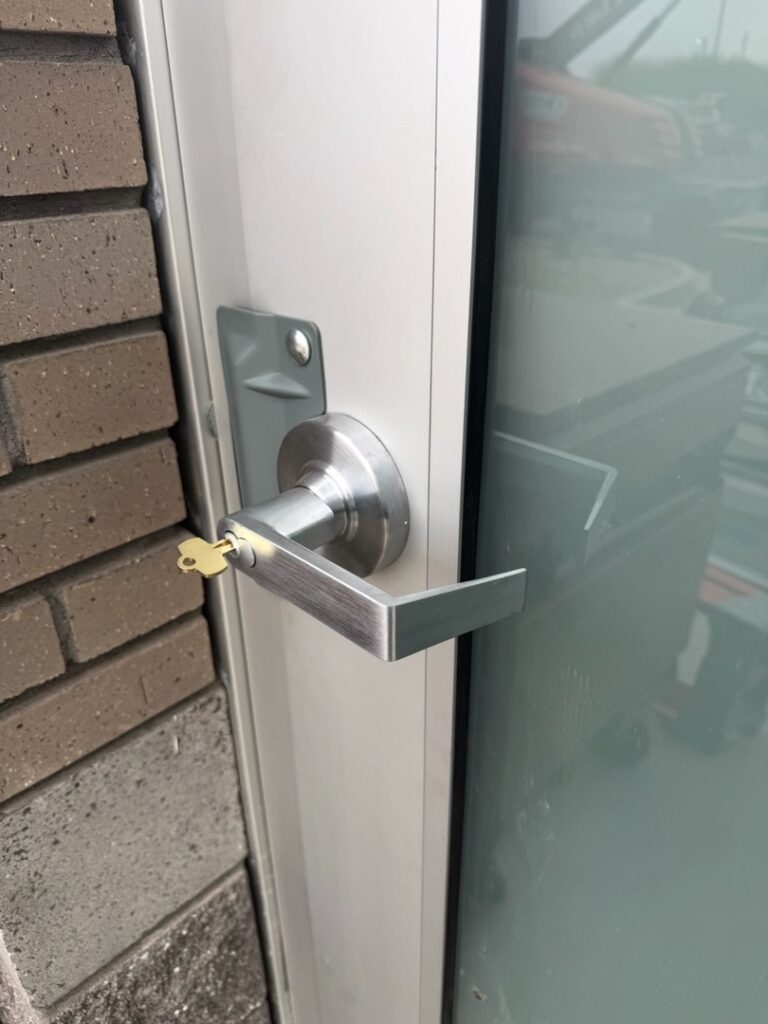 Commercial Door Lever Handle Lock Installation A brushed silver lever door handle and lock cylinder installed on a white aluminum commercial door frame next to a brick wall and glass panel.