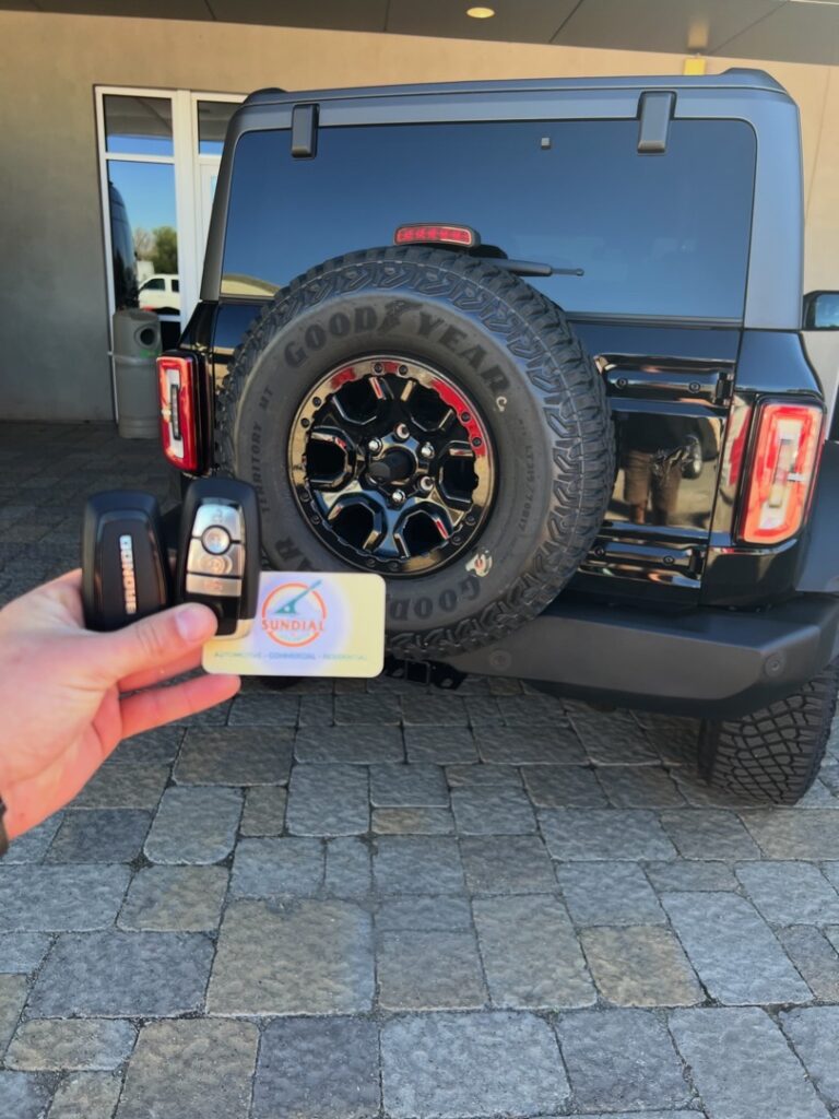 How Much Does a New Car Key Actually Cost Without a Spare? A hand holding a new key fob and smart key next to the rear spare tire of a dark blue Jeep Wrangler in a paved driveway.