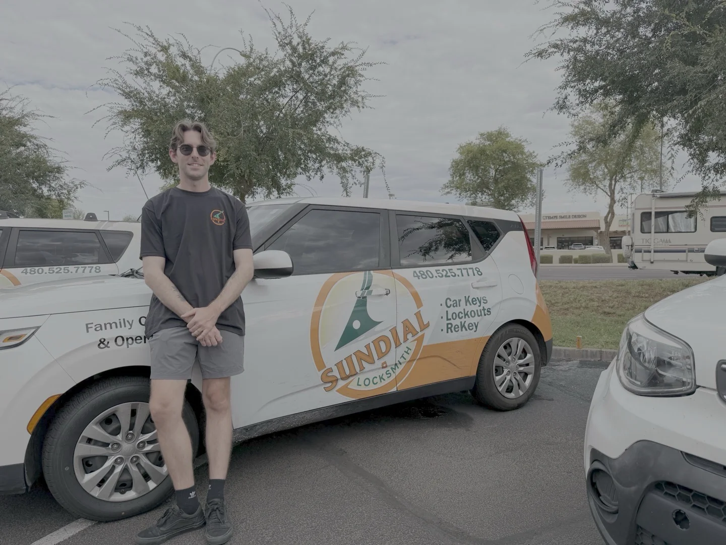 Door Viewer with Camera Installation: What to Expect and How It Works A young male Sundial Locksmith technician wearing sunglasses stands beside a white branded company car in an outdoor parking area.