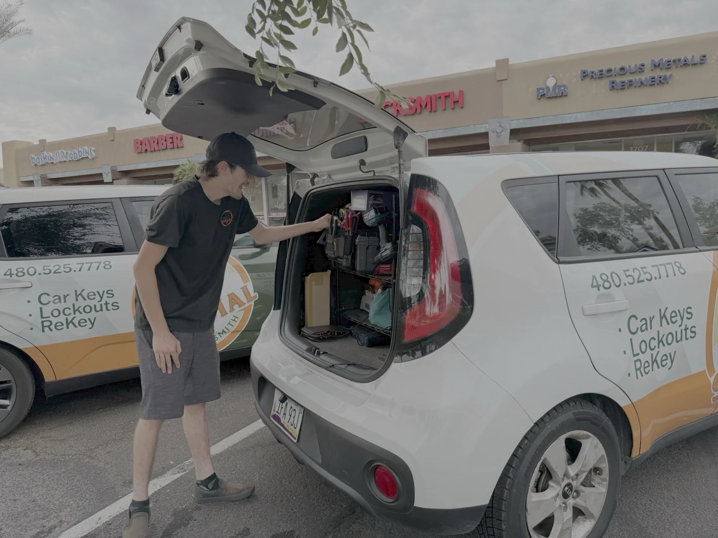 How Long Does 'All Keys Lost' Programming Actually Take? A locksmith technician in a black uniform opens the rear hatch of a branded white Kia Soul service vehicle stocked with tools and supplies in a strip mall parking lot.