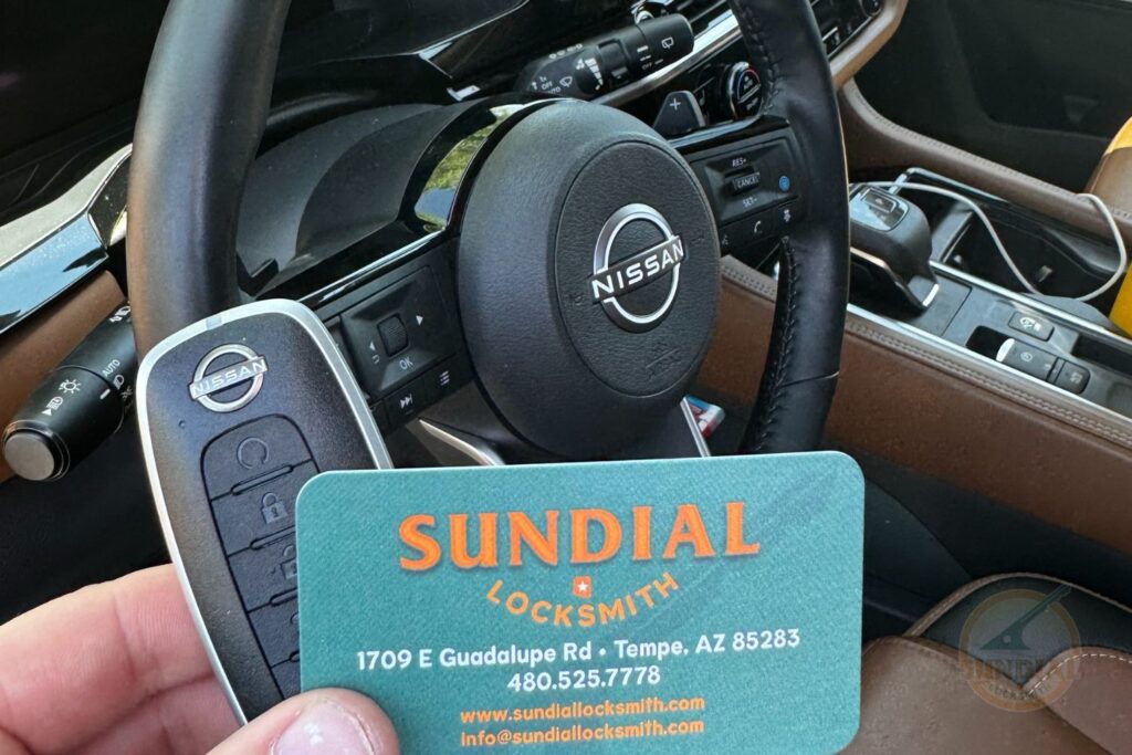 OEM Car Key Replacement: When It Matters and When It Doesn't A hand holding a Nissan smart key fob and a Sundial Locksmith business card inside a Nissan vehicle with the steering wheel visible in the background.