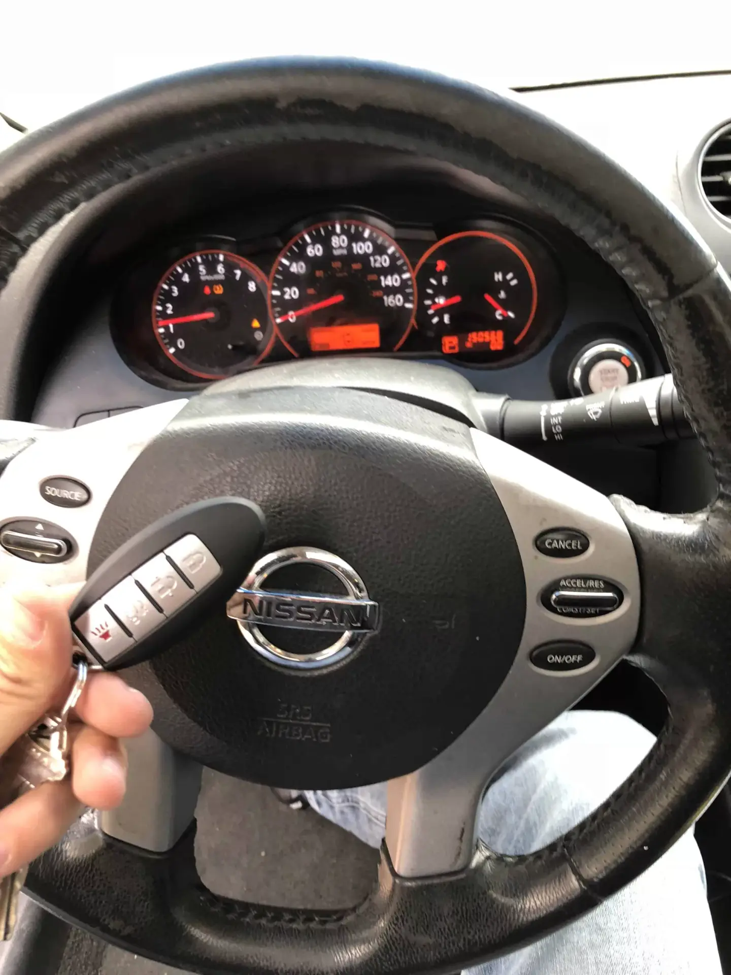 4 Things to Know If You Have Lost Your Car Keys A hand holding a Nissan smart key fob in front of a Nissan steering wheel with illuminated dashboard gauges visible in the background.